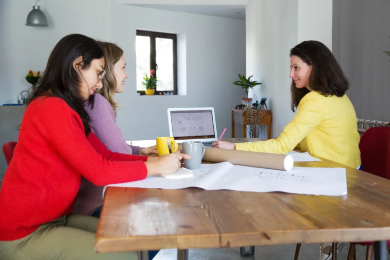 Three women sitting at a wooden table in an office, discussing architectural drawings with a laptop open in front of them.