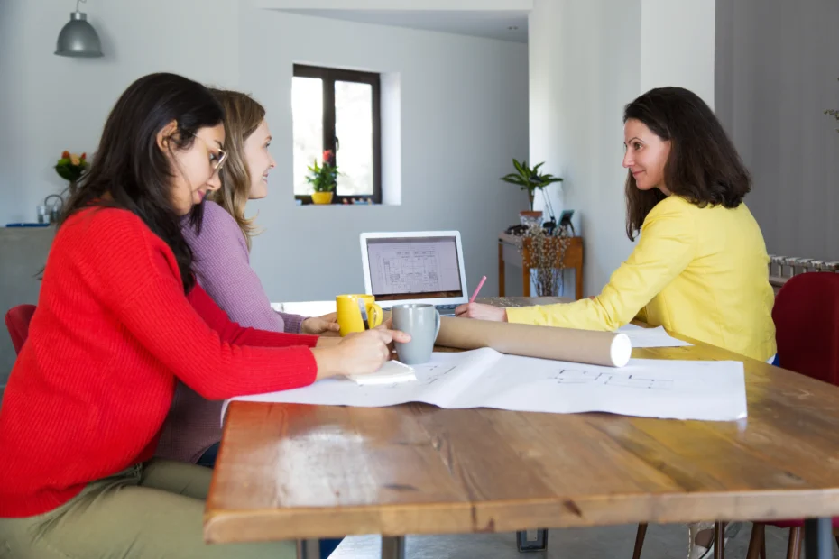 Three women sitting at a wooden table in an office, discussing architectural drawings with a laptop open in front of them.