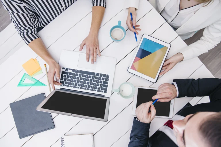 Top view of a group of coworkers working together at a white desk with a laptop, tablets, notebooks, coffee cups, and stationery items.