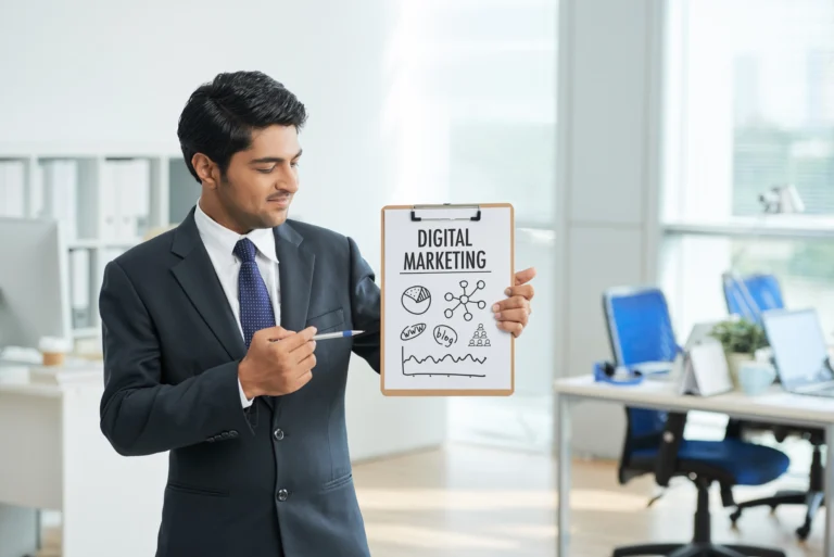 Businessman in a suit standing in an office, holding a clipboard with 'Digital Marketing' written on it and pointing to it with a pen.