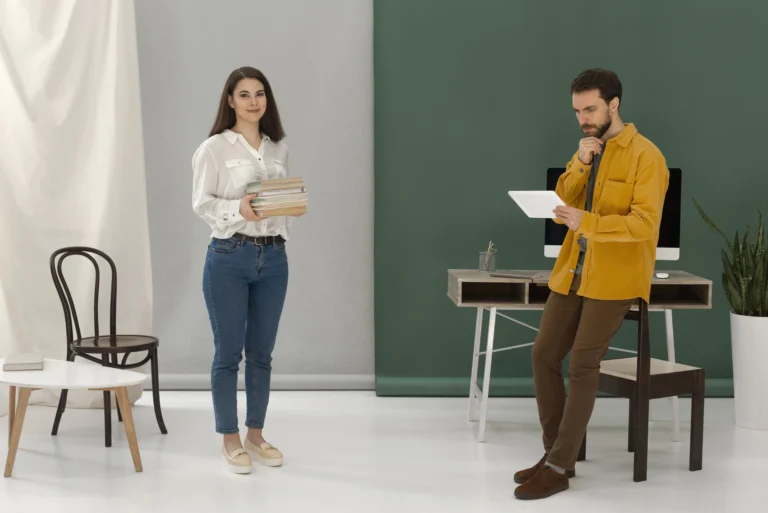 Woman holding a stack of books and smiling while a man stands near a desk using a tablet, in a modern minimal office setting.
